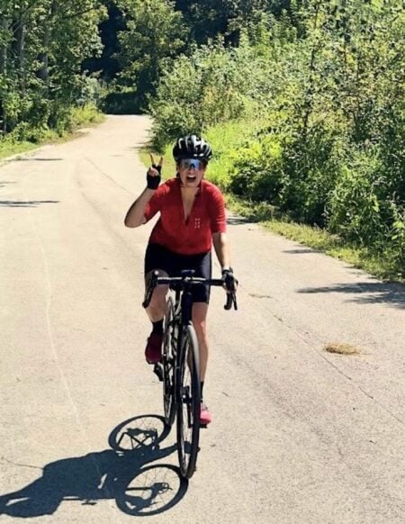 Une personne portant une chemise rouge et un casque noir profite du cyclotourisme Autriche en roulant à vélo sur un chemin pavé et bordé d'arbres, sous un soleil radieux. Le cycliste montre deux doigts en vivant l'aventure d'Autriche à vélo. - Ekilib