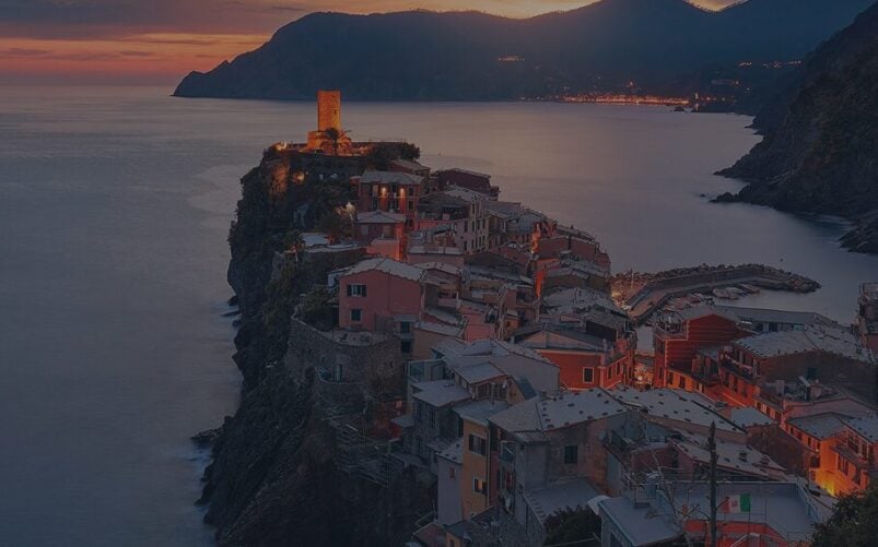 Un village côtier avec des bâtiments colorés et une tour se trouve au sommet des falaises surplombant la mer au coucher du soleil, éclairé par des lumières - un spectacle enchanteur le long du Grand Circuit de l'Empordà, avec les montagnes qui s'élèvent en arrière-plan. - Ekilib