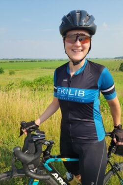 Une personne portant un casque de cycliste, des lunettes de soleil, des gants de cycliste et un maillot bleu et noir d'EKILIB se tient à côté d'un vélo dans un champ herbeux avec des terres agricoles et un ciel bleu en arrière-plan. - Ekilib
