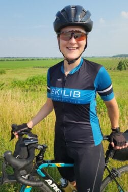Une personne portant un casque de cycliste, des lunettes de soleil, des gants de cycliste et un maillot bleu et noir d'EKILIB se tient à côté d'un vélo dans un champ herbeux avec des terres agricoles et un ciel bleu en arrière-plan. - Ekilib