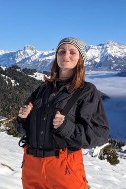 Flore Boubila se tient sur le versant d'une montagne enneigée, vêtue d'un bonnet rayé, d'une veste noire et d'un pantalon orange. Elle est entourée d'arbres et de montagnes enneigés, sous un ciel bleu clair, alors que la vallée est plongée dans le brouillard. - Ekilib