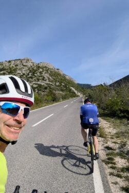 Deux cyclistes montent une route pavée entourée de collines et de verdure sous un ciel bleu clair. Francis Laporte, avec un casque blanc et des lunettes de soleil, sourit à la caméra tandis que l'autre cycliste pédale devant lui. - Ekilib