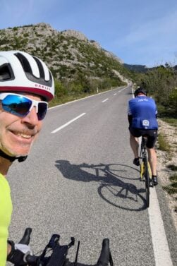 Deux cyclistes montent une route pavée entourée de collines et de verdure sous un ciel bleu clair. Francis Laporte, avec un casque blanc et des lunettes de soleil, sourit à la caméra tandis que l'autre cycliste pédale devant lui. - Ekilib