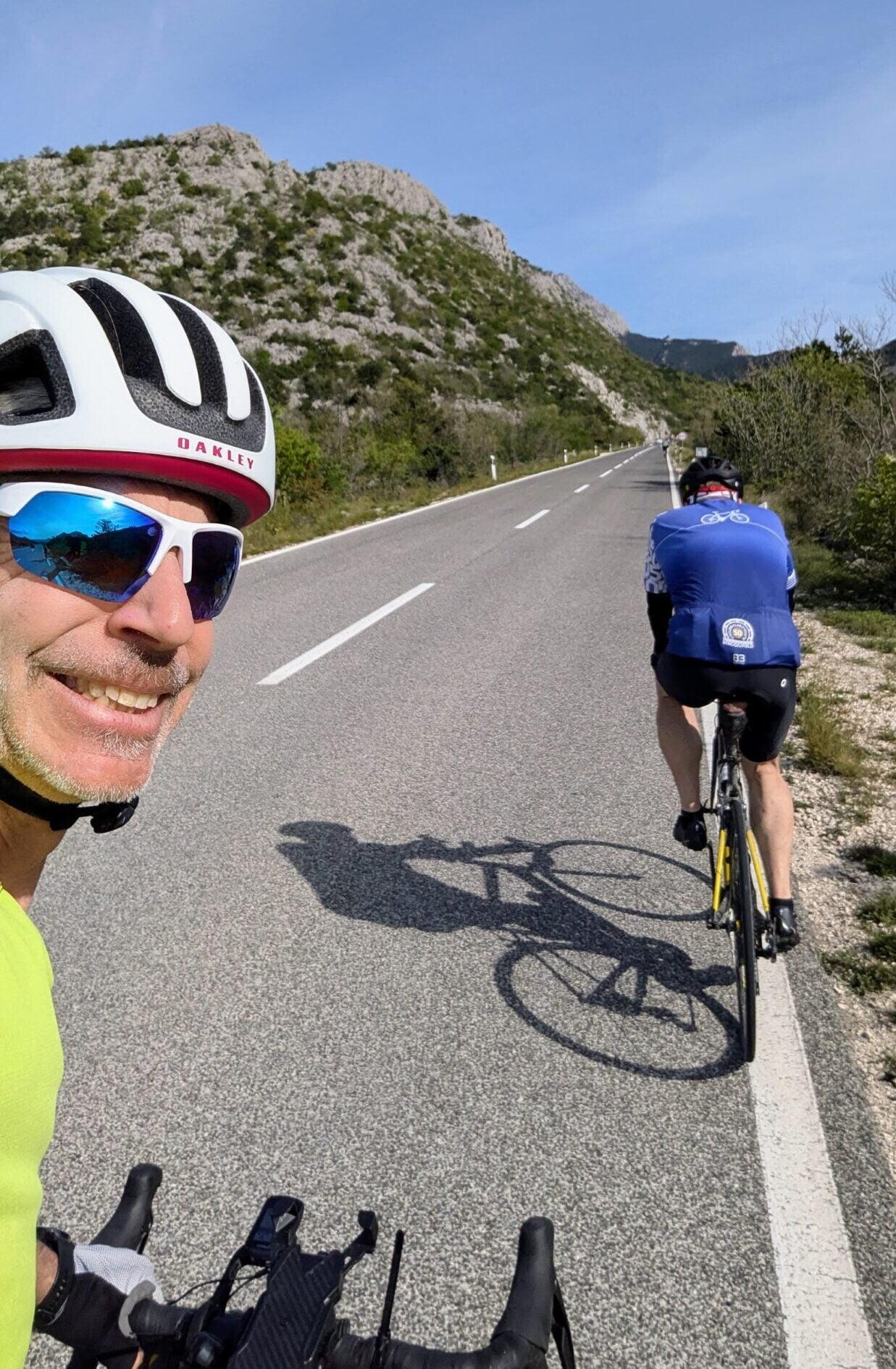 Deux cyclistes montent une route pavée entourée de collines et de verdure sous un ciel bleu clair. Francis Laporte, avec un casque blanc et des lunettes de soleil, sourit à la caméra tandis que l'autre cycliste pédale devant lui. - Ekilib