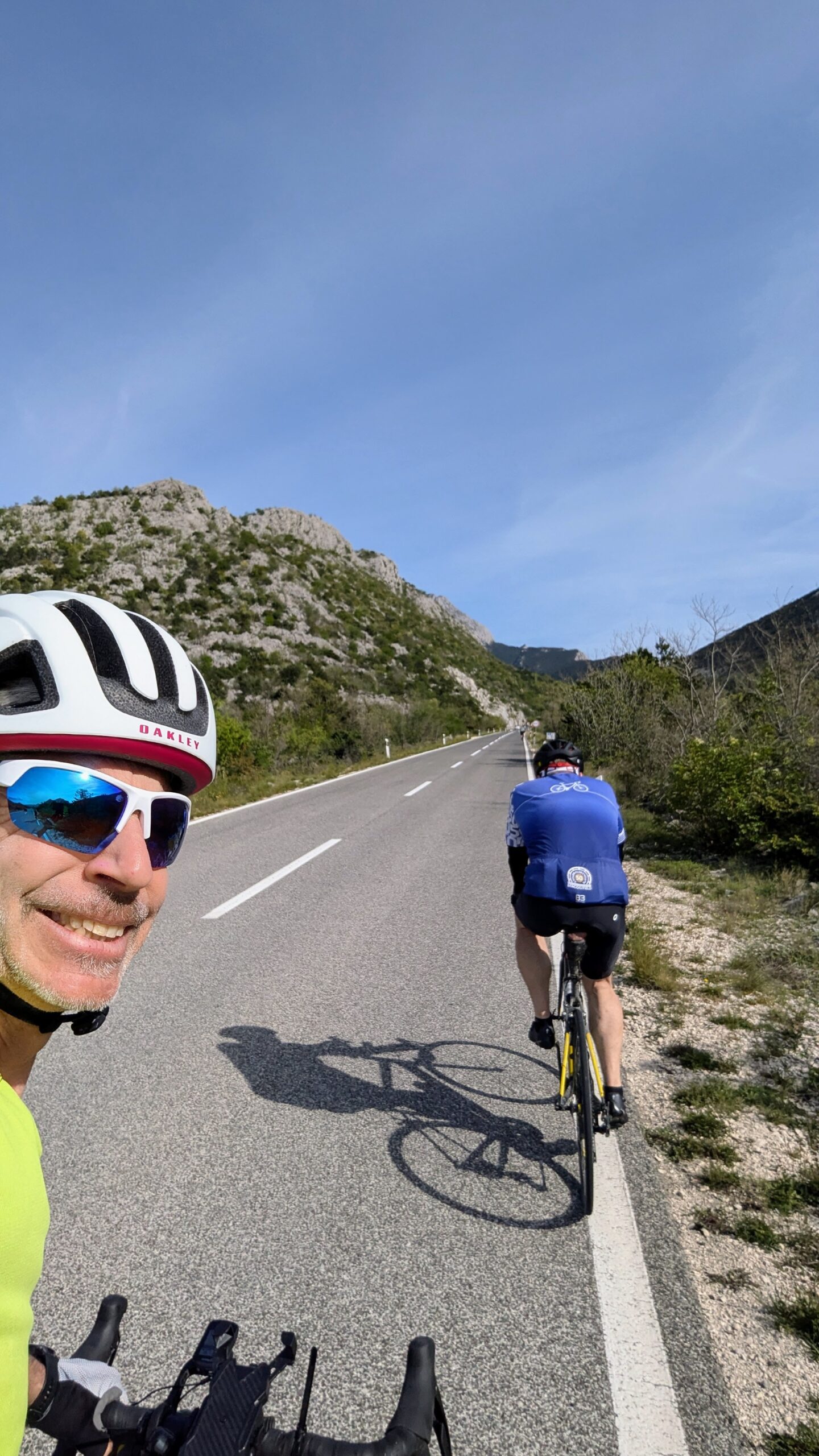 Deux cyclistes montent une route pavée entourée de collines et de verdure sous un ciel bleu clair. Francis Laporte, avec un casque blanc et des lunettes de soleil, sourit à la caméra tandis que l'autre cycliste pédale devant lui. - Ekilib