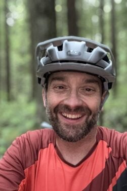 François-Xavier Gagnon, portant un casque de vélo gris et un maillot de sport orange, sourit à l'appareil photo dans une zone forestière avec des arbres et de la verdure en arrière-plan flou. - Ekilib