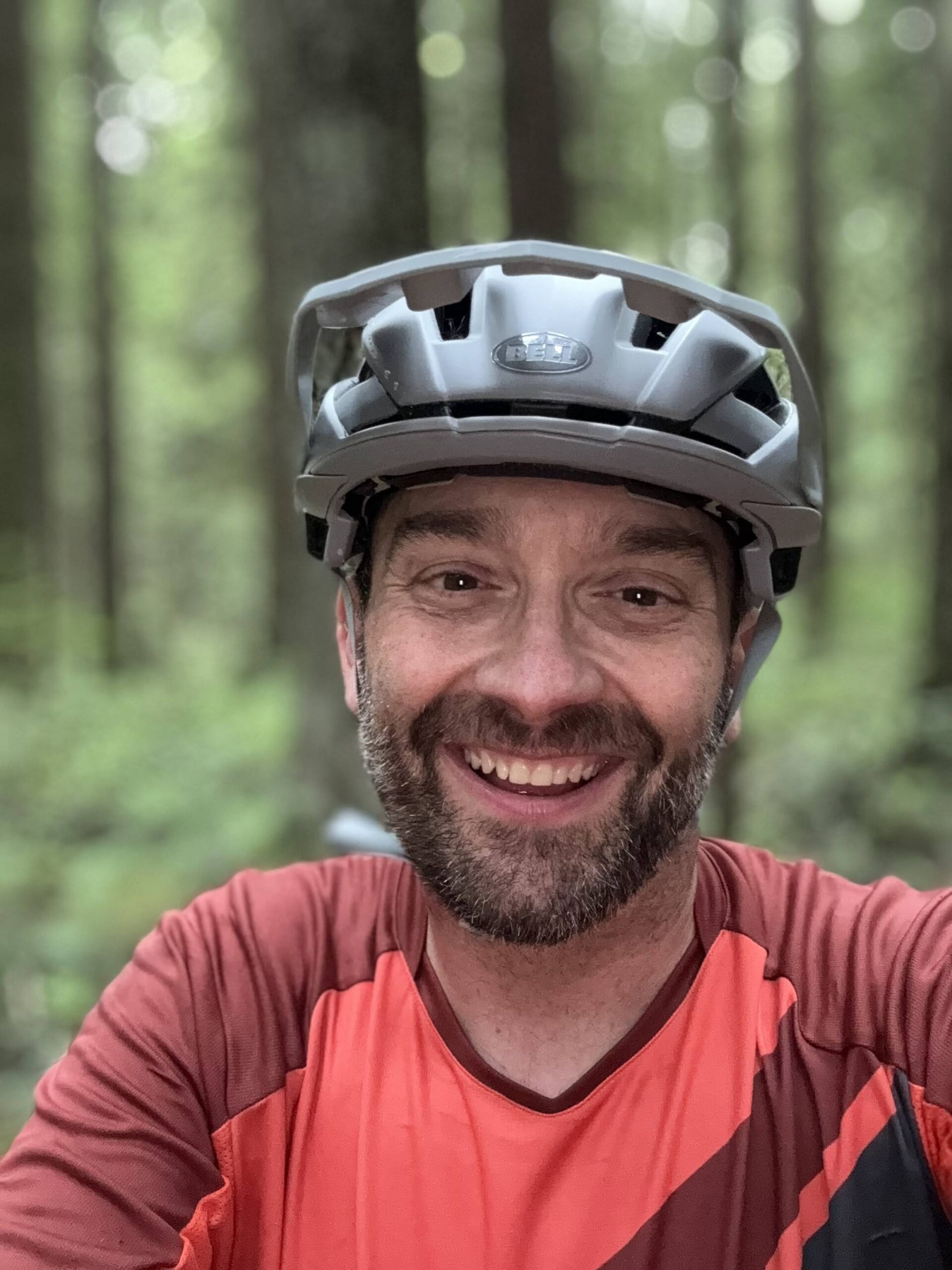 François-Xavier Gagnon, portant un casque de vélo gris et un maillot de sport orange, sourit à l'appareil photo dans une zone forestière avec des arbres et de la verdure en arrière-plan flou. - Ekilib