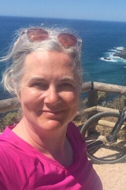 Hélène Lauzière, cheveux gris et lunettes de soleil sur la tête, prend un selfie près d'une barrière en bois donnant sur l'océan et la côte rocheuse. Un vélo est partiellement visible à droite. - Ekilib