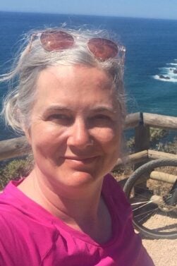 Hélène Lauzière, cheveux gris et lunettes de soleil sur la tête, prend un selfie près d'une barrière en bois donnant sur l'océan et la côte rocheuse. Un vélo est partiellement visible à droite. - Ekilib