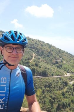 Jean-François Gagné, vêtu d'un maillot cycliste bleu, d'un casque et de lunettes, se tient devant une balustrade en bois avec des collines verdoyantes et une route sinueuse en arrière-plan, par une journée ensoleillée. - Ekilib