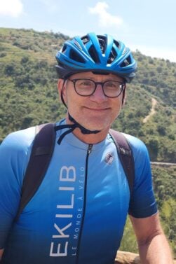 Jean-François Gagné, vêtu d'un maillot cycliste bleu, d'un casque et de lunettes, se tient devant une balustrade en bois avec des collines verdoyantes et une route sinueuse en arrière-plan, par une journée ensoleillée. - Ekilib