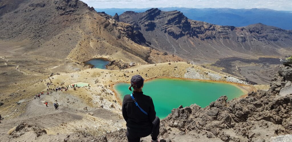 Une personne portant une veste et une casquette noires est assise sur une corniche rocheuse surplombant deux lacs d'un turquoise éclatant dans des montagnes arides, avec des groupes de randonneurs en contrebas - un paysage majestueux que Lorène Berizzi recherche souvent dans ses aventures. - Ekilib