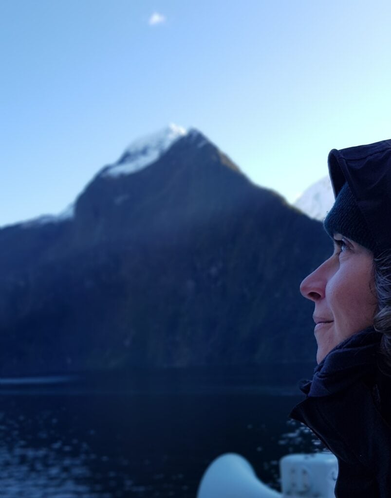 Une personne vêtue d'une veste sombre et d'un bonnet est représentée de profil sur le côté droit de l'image, avec derrière elle une montagne aux sommets enneigés et un plan d'eau flous sous un ciel clair, évoquant l'esprit d'aventure de Lorène Berizzi. - Ekilib