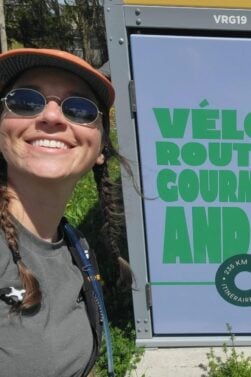 Lorène Berizzi, lunettes de soleil, casquette et tresses, sourit à côté d'un panneau indiquant Vélo route gourm avec une étiquette circulaire indiquant 235 KM - Itinéraire Gourm. L'arrière-plan est couvert d'une végétation luxuriante. - Ekilib