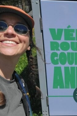 Lorène Berizzi, lunettes de soleil, casquette et tresses, sourit à côté d'un panneau indiquant Vélo route gourm avec une étiquette circulaire indiquant 235 KM - Itinéraire Gourm. L'arrière-plan est couvert d'une végétation luxuriante. - Ekilib