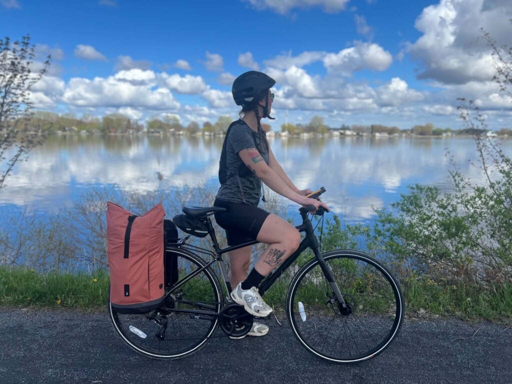 Lorène Berizzi, coiffée d'un casque et vêtue d'une tenue de cycliste, roule sur un vélo avec une grande sacoche rose au bord d'un lac calme, sous un ciel partiellement nuageux. Des arbres et de la verdure bordent l'eau. - Ekilib