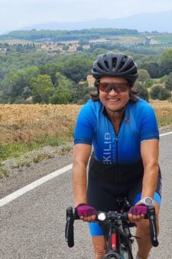 Une personne portant une tenue de cycliste bleue, des gants, des lunettes de soleil et un casque roule à vélo sur une route de campagne avec des champs vallonnés et des arbres en arrière-plan. - Ekilib