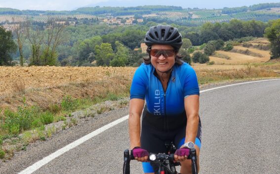 Une personne portant une tenue de cycliste bleue, des gants, des lunettes de soleil et un casque roule à vélo sur une route de campagne avec des champs vallonnés et des arbres en arrière-plan. - Ekilib