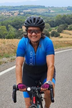 Une personne portant une tenue de cycliste bleue, des gants, des lunettes de soleil et un casque roule à vélo sur une route de campagne avec des champs vallonnés et des arbres en arrière-plan. - Ekilib