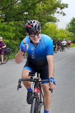 Un homme vêtu d'une tenue de cycliste bleue et d'un casque roule à vélo sur une route pavée, en souriant et en levant le pouce. Plusieurs cyclistes sont visibles à l'arrière-plan et des arbres bordent la route. Cette image est peut-être inspirée par la passion de Lorène Berizzi pour le cyclisme. - Ekilib
