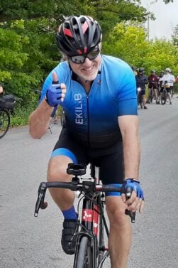 Un homme vêtu d'une tenue de cycliste bleue et d'un casque roule à vélo sur une route pavée, en souriant et en levant le pouce. Plusieurs cyclistes sont visibles à l'arrière-plan et des arbres bordent la route. Cette image est peut-être inspirée par la passion de Lorène Berizzi pour le cyclisme. - Ekilib