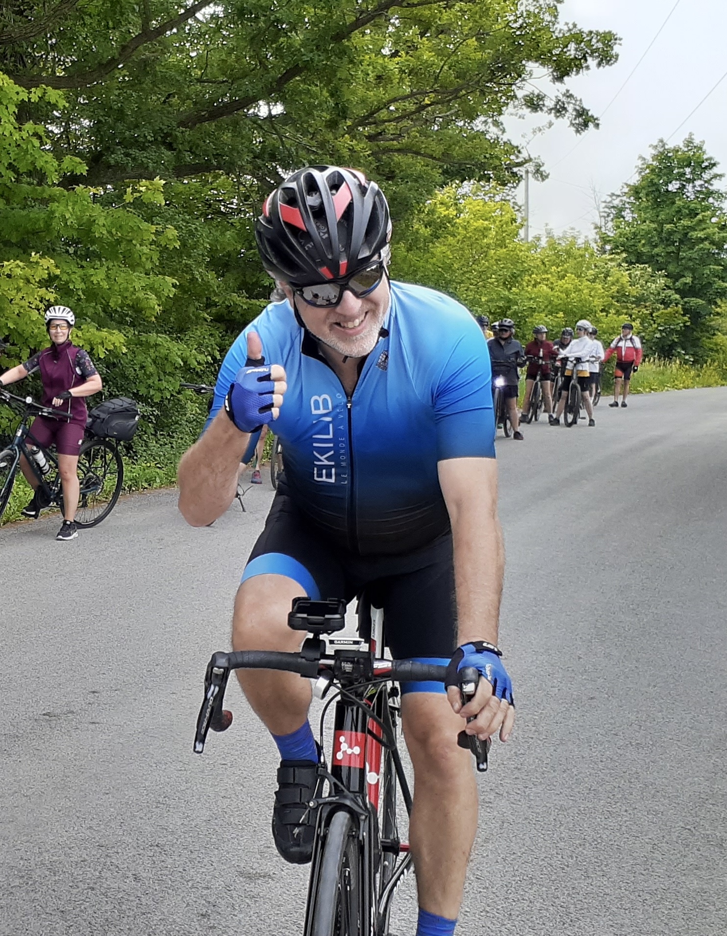 Un homme vêtu d'une tenue de cycliste bleue et d'un casque roule à vélo sur une route pavée, en souriant et en levant le pouce. Plusieurs cyclistes sont visibles à l'arrière-plan et des arbres bordent la route. Cette image est peut-être inspirée par la passion de Lorène Berizzi pour le cyclisme. - Ekilib