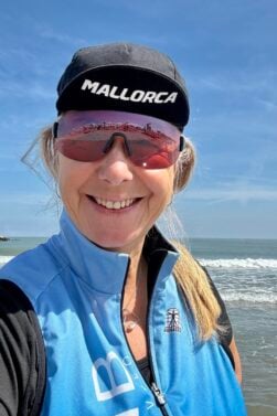 Sonia Laverdière, portant une casquette de Majorque, des lunettes de soleil réfléchissantes et une veste sans manche bleue, sourit à l'appareil photo alors qu'elle se tient sur une plage avec des vagues et un ciel bleu en arrière-plan. - Ekilib