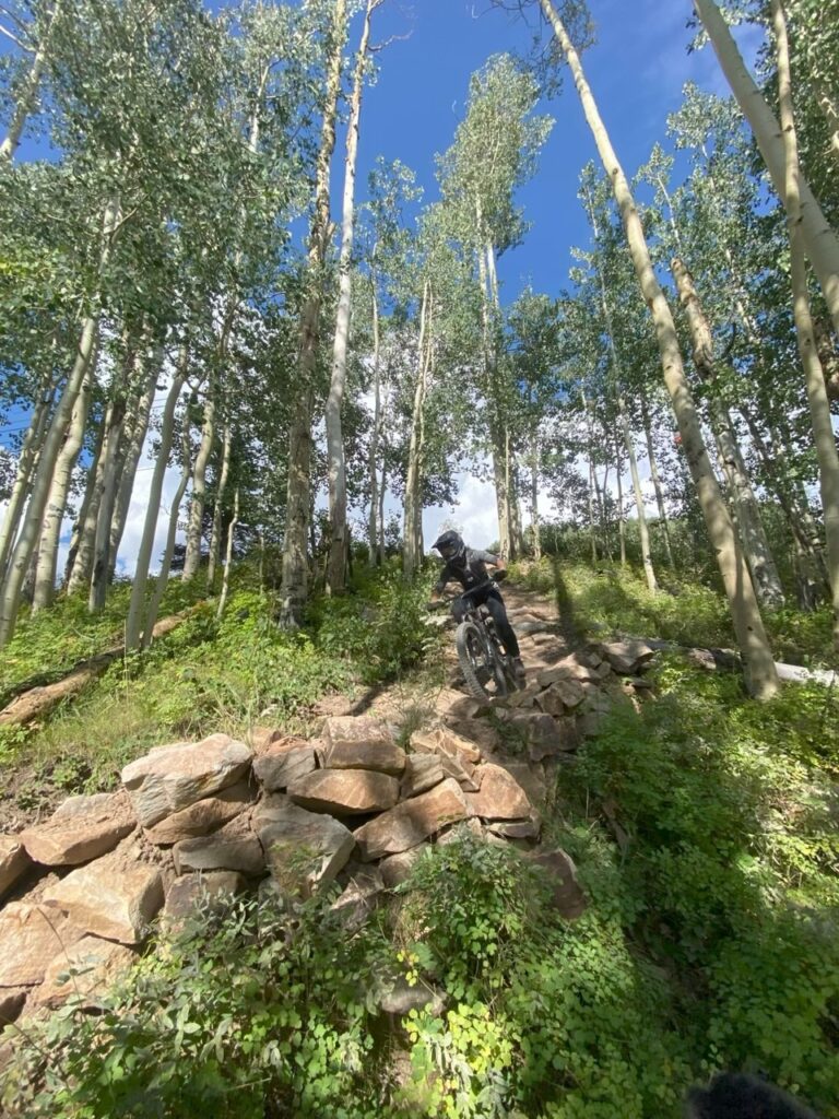 Valérie Beauregard, coiffée d'un casque, descend en VTT un sentier rocailleux dans une forêt aux arbres hauts et fins, sous un ciel bleu parsemé de nuages. - Ekilib