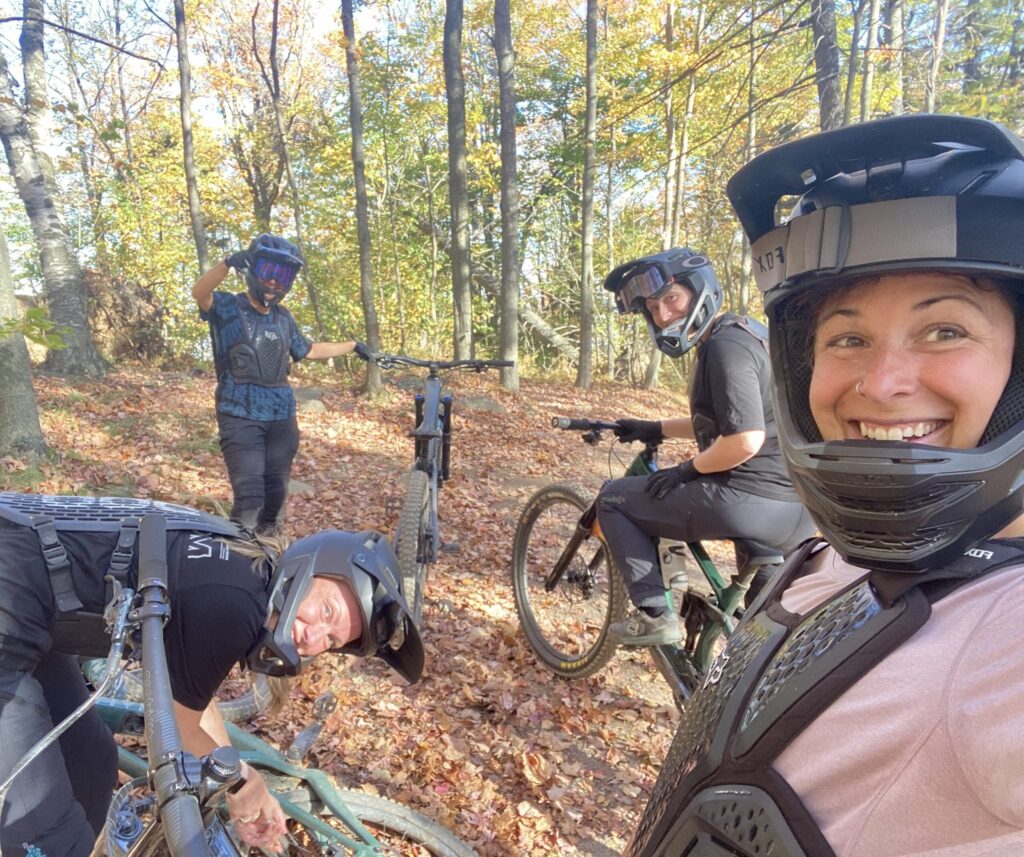 Quatre personnes, dont Valérie Beauregard, portant des équipements de protection et des casques, posent avec des VTT dans une forêt aux feuilles d'automne. L'une d'entre elles sourit à l'appareil photo, tandis qu'une autre s'accroupit près d'une roue de vélo. - Ekilib