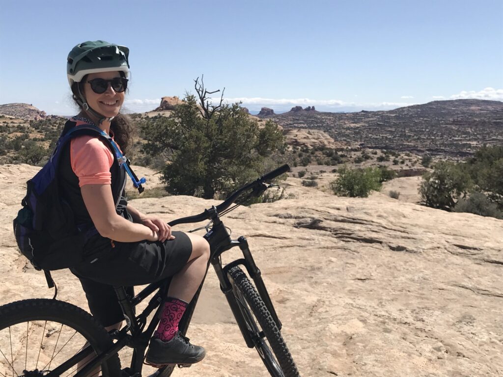 Valérie Beauregard, portant un casque, des lunettes de soleil et un sac à dos, est assise sur un VTT sur un sentier rocailleux avec un paysage désertique, des buissons épars et des formations rocheuses lointaines sous un ciel clair. - Ekilib