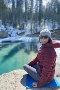 Valérie Beauregard, vêtue d'un manteau rouge, d'un bonnet gris et de lunettes de soleil, est assise sur un rocher au bord d'une rivière claire et partiellement gelée, avec de la neige et des arbres à feuilles persistantes en arrière-plan. - Ekilib