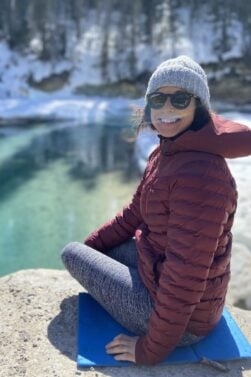 Valérie Beauregard, vêtue d'un manteau rouge, d'un bonnet gris et de lunettes de soleil, est assise sur un rocher au bord d'une rivière claire et partiellement gelée, avec de la neige et des arbres à feuilles persistantes en arrière-plan. - Ekilib