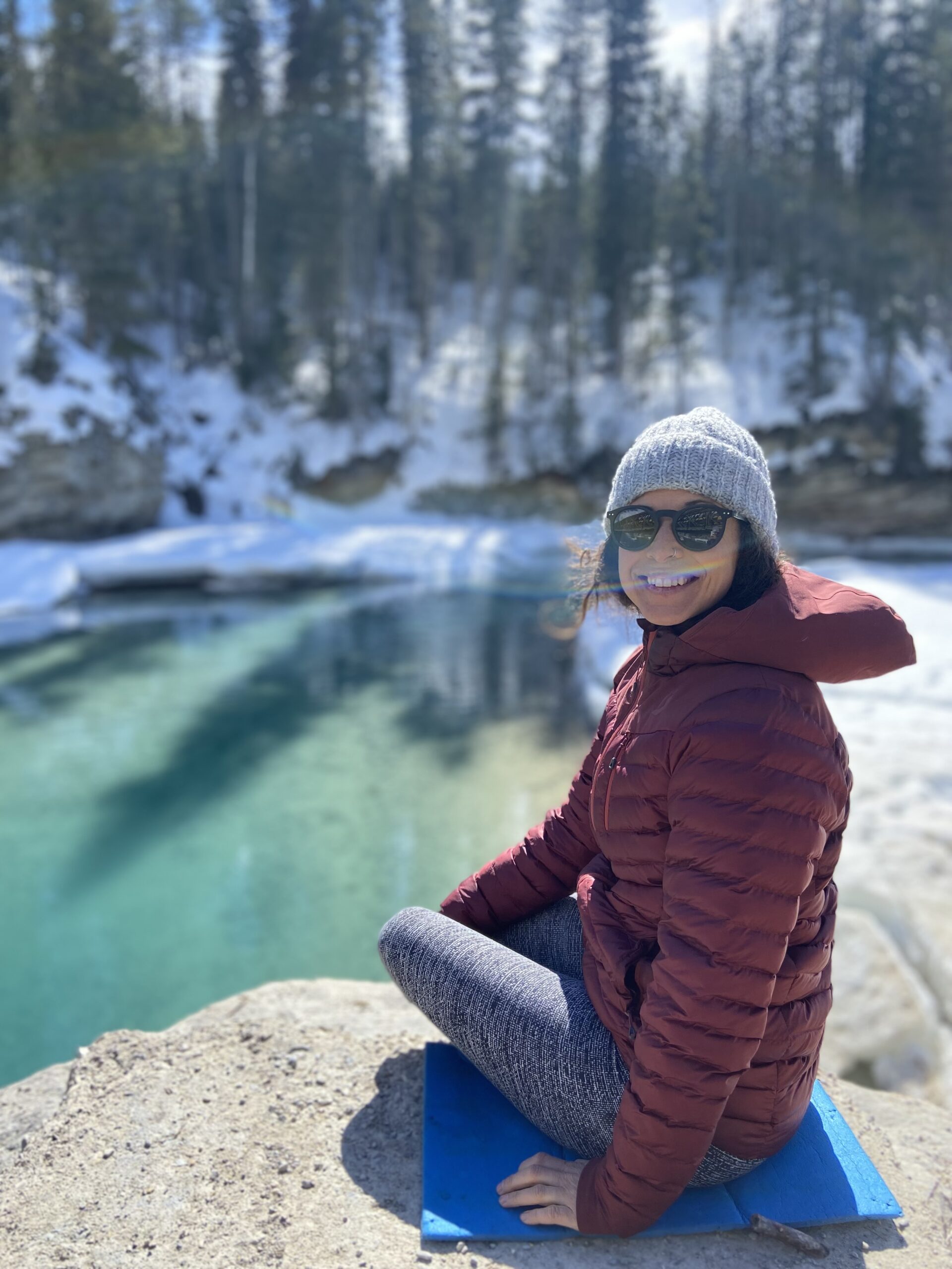 Valérie Beauregard, vêtue d'un manteau rouge, d'un bonnet gris et de lunettes de soleil, est assise sur un rocher au bord d'une rivière claire et partiellement gelée, avec de la neige et des arbres à feuilles persistantes en arrière-plan. - Ekilib