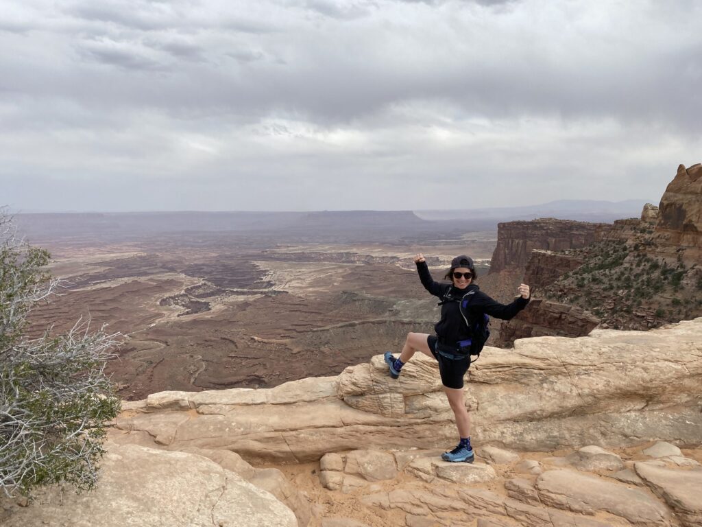 Valérie Beauregard, vêtue d'une tenue de sport noire et d'un sac à dos, se tient sur une corniche rocheuse, levant les bras en signe de triomphe, avec pour toile de fond un vaste paysage de canyon et un ciel nuageux. - Ekilib