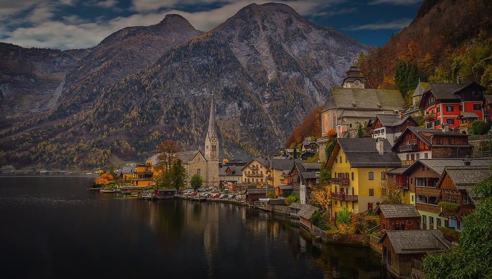 Un village au bord d'un lac, le long de la Route des lacs Autriche, avec des maisons colorées et une flèche d'église, se trouve au pied de montagnes boisées. Un ciel nuageux et un feuillage d'automne vibrant dans les tons orange et jaune complètent la scène pittoresque. - Ekilib