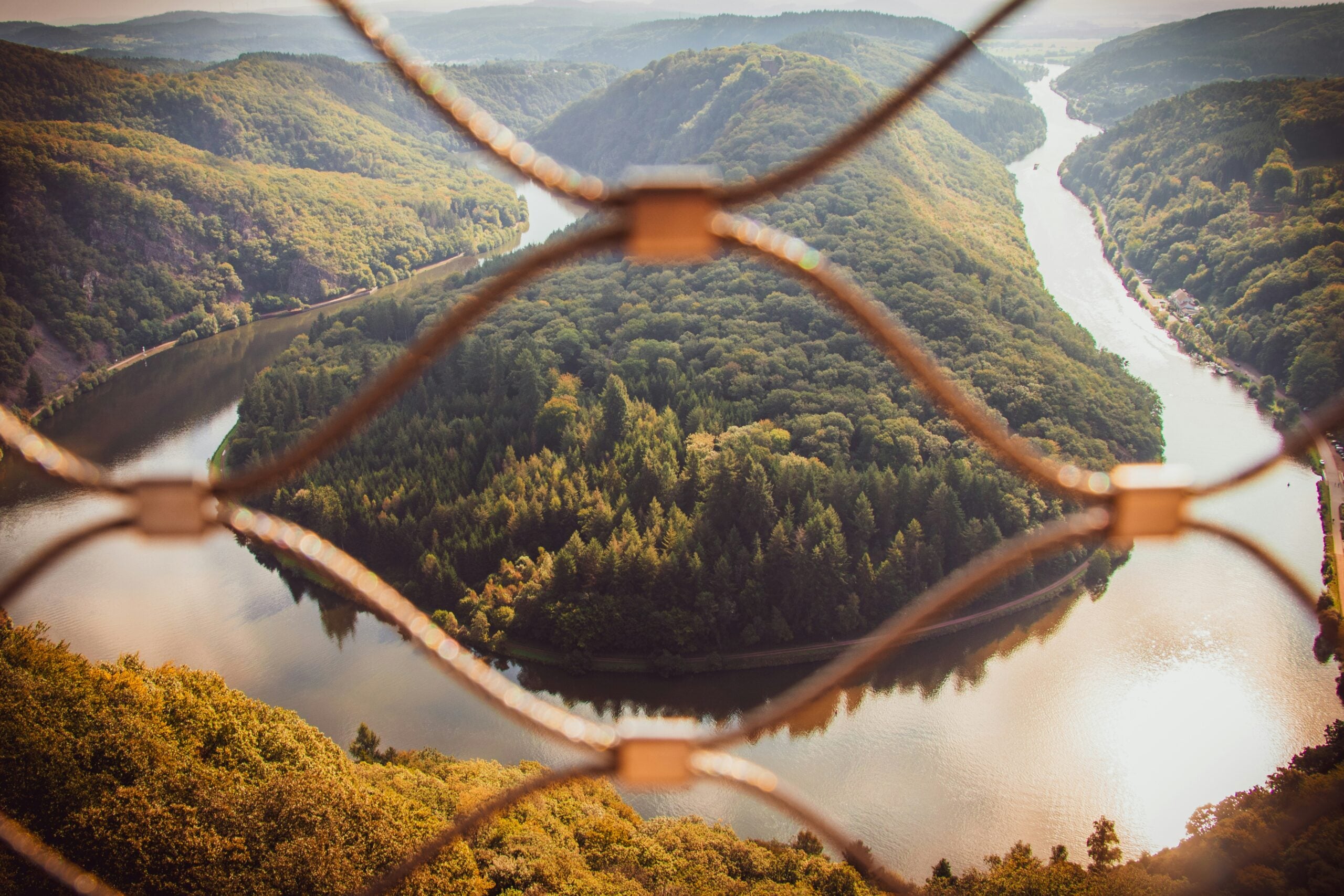Un coude de rivière couvert d'arbres est vu d'en haut à travers une clôture métallique, avec des collines et des forêts qui s'étendent au loin sous un ciel brumeux. - Ekilib