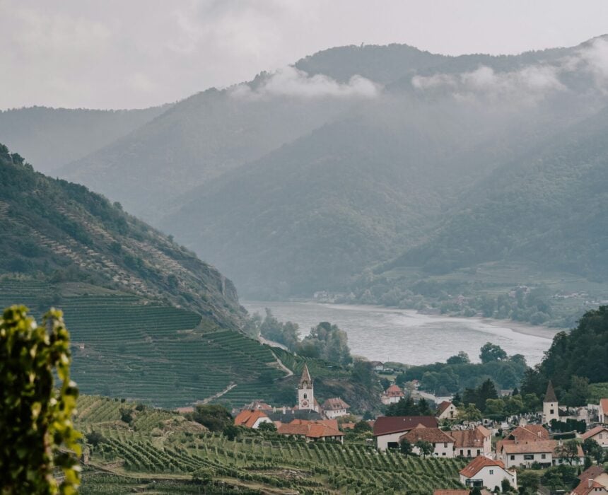 Un village aux maisons à toit rouge et à l'église se trouve au milieu de vignobles verdoyants le long du Danube, et vous invite à découvrir son charme lors d'un voyage à vélo, avec un fleuve et des montagnes boisées sous un ciel nuageux. - Ekilib