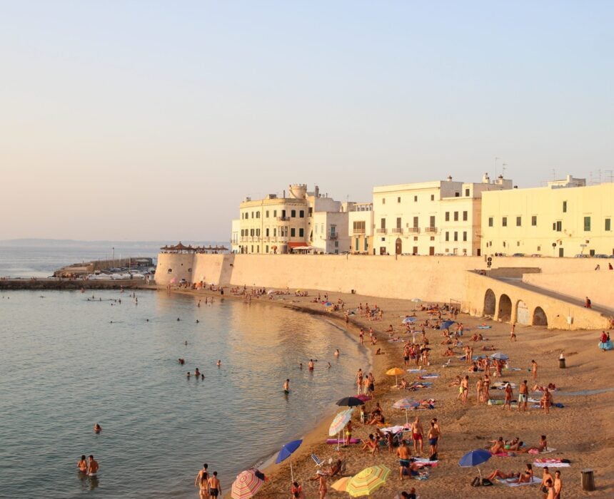 Des gens prennent des bains de soleil et nagent sur une plage de sable le long de la ville côtière fortifiée de Gallipoli, aux bâtiments blancs. La mer est calme et le ciel est dégagé, reflétant l'esprit de liberté de cette journée chaude et ensoleillée. - Ekilib