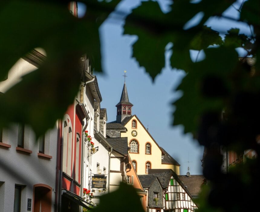 Vue d'un village-rue européen le long de la Route des Vins d'Alsace, avec des bâtiments colorés et un clocher d'église central en arrière-plan, partiellement encadrés par des feuilles vertes et des ombres au premier plan. - Ekilib