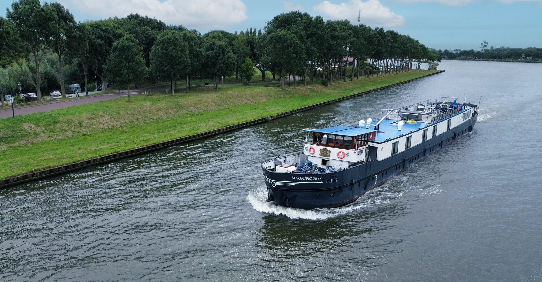 Une péniche fluviale noire et blanche nommée Magnifique II navigue le long d'un canal bordé d'une digue herbeuse et d'une rangée d'arbres par une journée nuageuse. - Ekilib