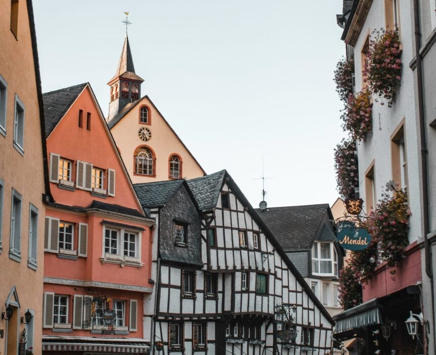 Une rue pavée étroite bordée de bâtiments colorés et d'une maison à colombages ; des tables et des chaises sont installées à l'extérieur, et une église avec une haute flèche se dresse à l'arrière-plan - parfait pour explorer à vélo la charmante région de Moselle-Sarre sous un ciel dégagé. - Ekilib