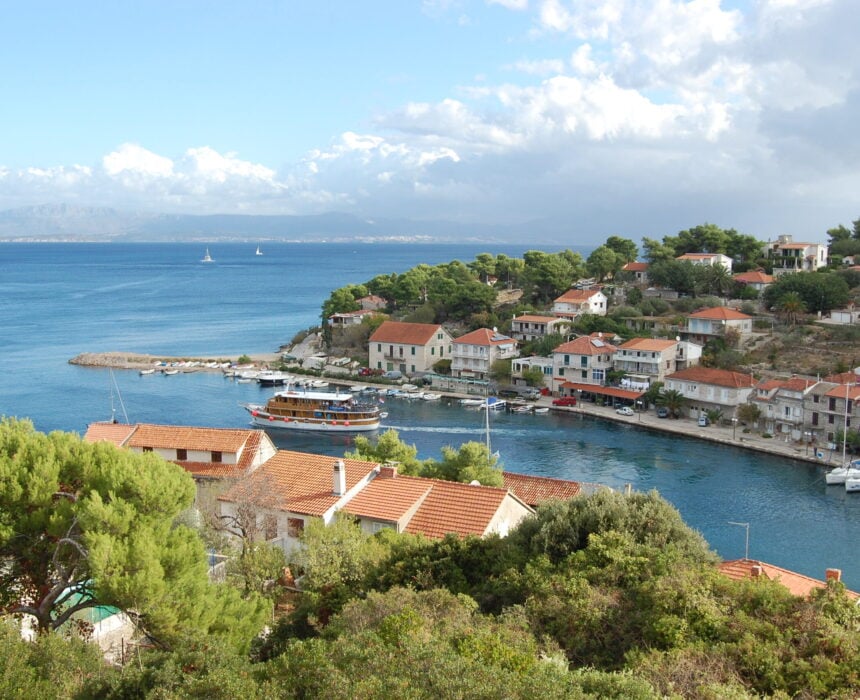 Village côtier avec des maisons aux toits rouges près d'un port bleu, de petits bateaux et un plus grand bateau en bois à quai - parfait pour explorer la Croatie à vélo-bateau - avec des arbres verts au premier plan et des collines lointaines de l'autre côté de la mer sous un ciel partiellement nuageux. - Ekilib