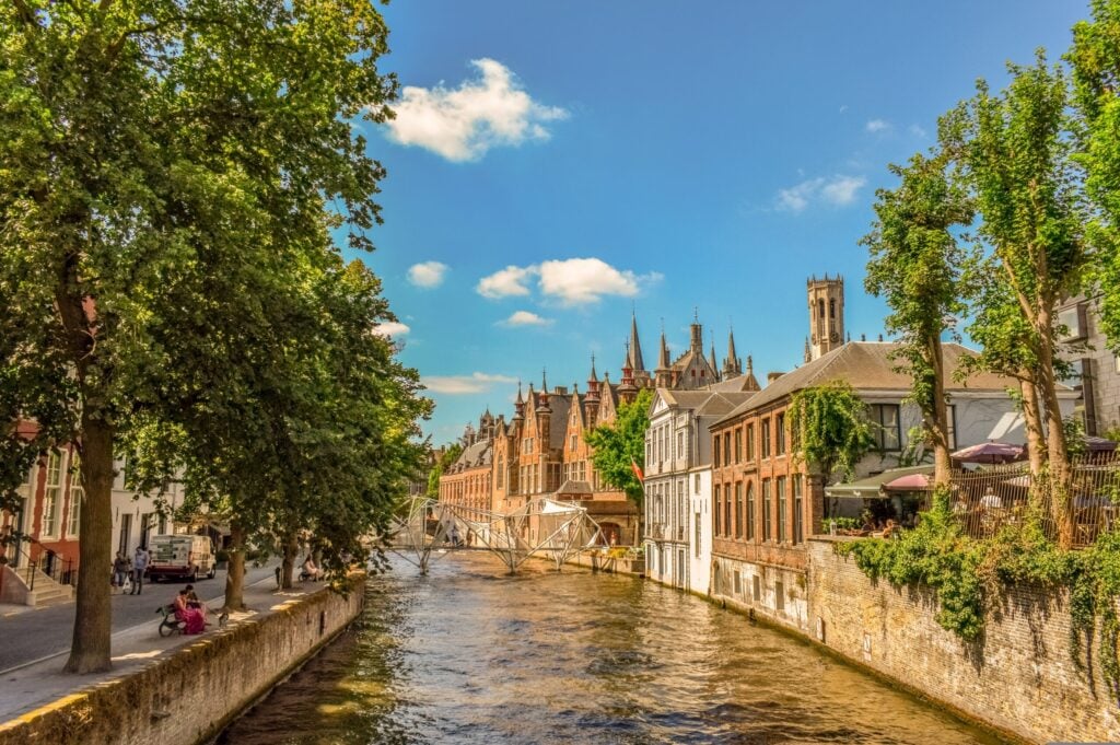 Un canal traverse une ville bordée de bâtiments historiques en briques et d'arbres, sous un ciel bleu parsemé de nuages. Des gens se promènent et s'assoient le long du canal, et un pont est visible au loin. - Ekilib