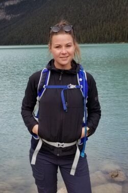 Cécile Bellissard, vêtue d'une veste noire et d'un sac à dos, se tient au bord d'un lac turquoise encadré de montagnes et de pins, sous un ciel partiellement nuageux. - Ekilib