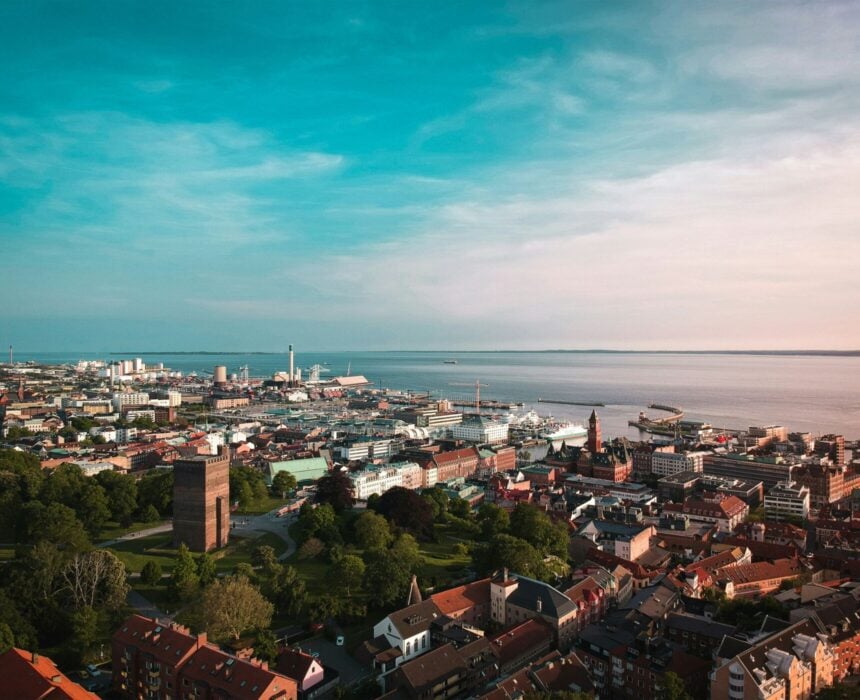 Vue aérienne d'une ville côtière le long de la route Kattegattleden, avec des bâtiments aux toits rouges, des arbres verts, des structures industrielles au bord de l'eau et une grande étendue d'eau s'étendant jusqu'à l'horizon, sous un ciel partiellement nuageux. - Ekilib