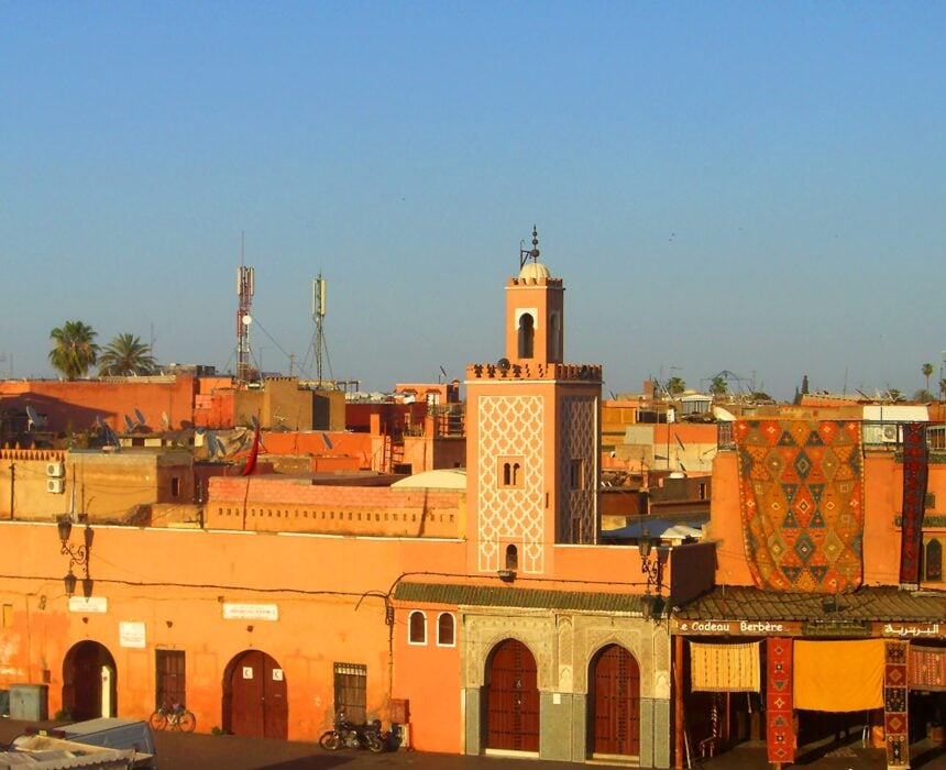 Un paysage urbain composé de bâtiments brun-rouge, d'une grande mosquée avec un minaret à motifs et d'un ciel bleu clair. Des antennes et des palmiers sont visibles à l'arrière-plan. La scène suggère un cadre nord-africain ou moyen-oriental. - Ekilib