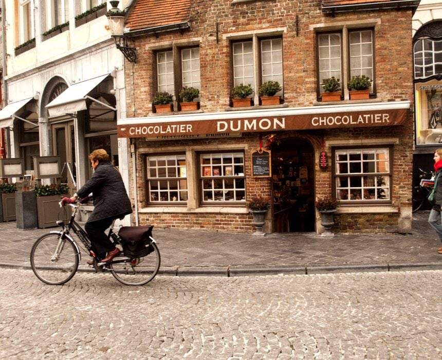 Une personne passe à vélo devant Dumon, un magasin de chocolats en briques avec une enseigne brune, des plantes sur les rebords des fenêtres et de grandes vitrines dans une rue pavée. Deux autres personnes marchent à proximité. - Ekilib