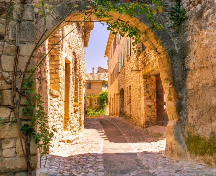 Une rue pavée étroite dans un village historique, vue à travers une vieille arche en pierre, avec des bâtiments en pierre et un peu de verdure baignés de soleil - parfait pour une promenade en vélo en Provence sous un ciel clair et ensoleillé. - Ekilib