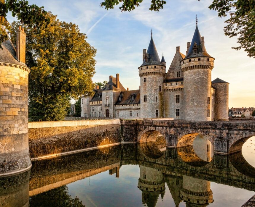 Un château en pierre avec des tours pointues et un pont traverse une douve, se reflétant dans l'eau calme. Le soleil jette une lumière chaude sur le bâtiment et les arbres qui l'entourent. Une étape enchanteresse sur la route de la vallée de la Loire à vélo. - Ekilib