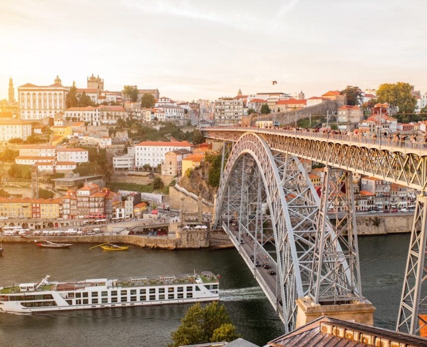 Un grand pont métallique en arc traverse une rivière et un bateau de croisière fluviale blanc passe en dessous. Des bâtiments colorés couvrent le flanc de la colline à l'arrière-plan, baigné par la lumière du soleil - une vue panoramique souvent admirée sur la route du Minho à vélo Portugal. - Ekilib